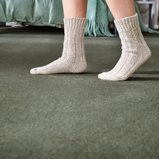 Detailaufnahme von hellen Stricksocken auf grünem JOKA Teppichboden im Schlafzimmer.