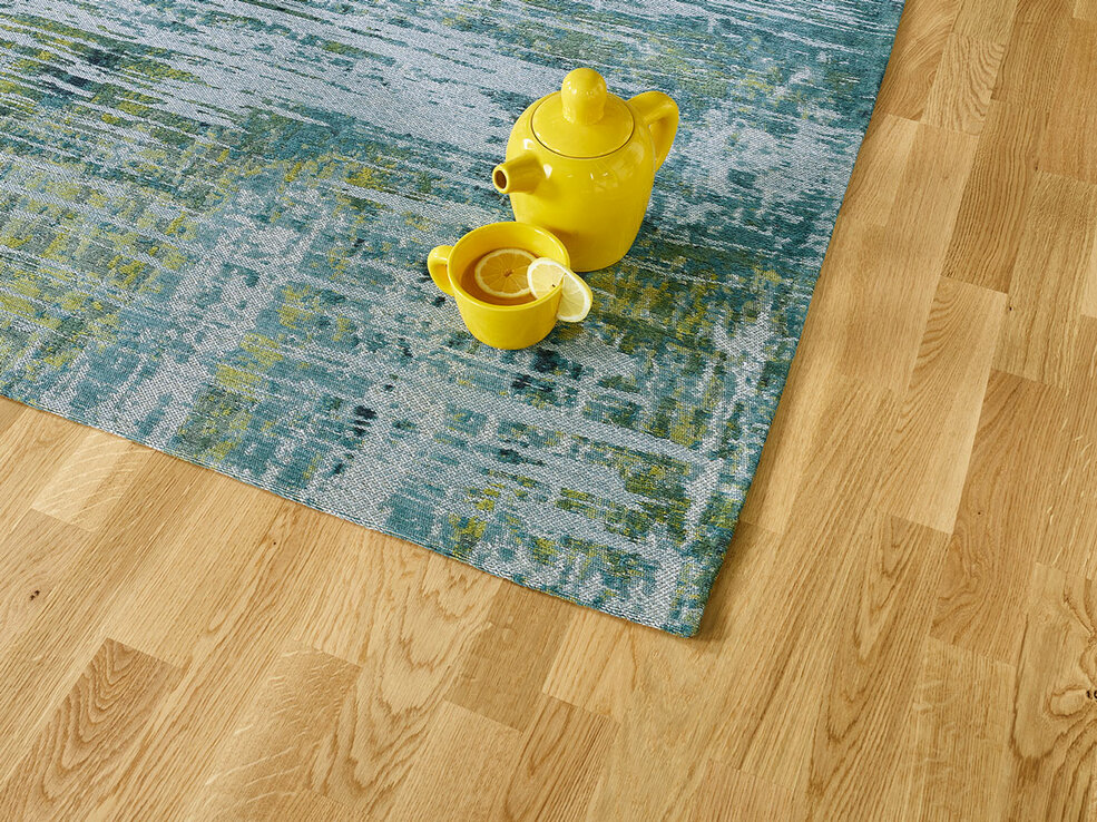 Nahaufnahme eines natürlichen JOKA Parkettbodens. Auf dem Fußboden liegt ein blau gelb gemusterter JOKA Teppich.