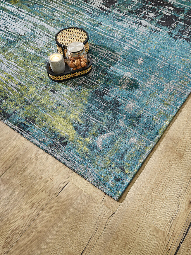 Nahaufnahme eines Eiche Natur JOKA Laminats. Auf dem Boden liegt ein blauer JOKA Teppich mit Mustern in unterschiedlichen Farben, passend zur Grünanlage Wohnidee.