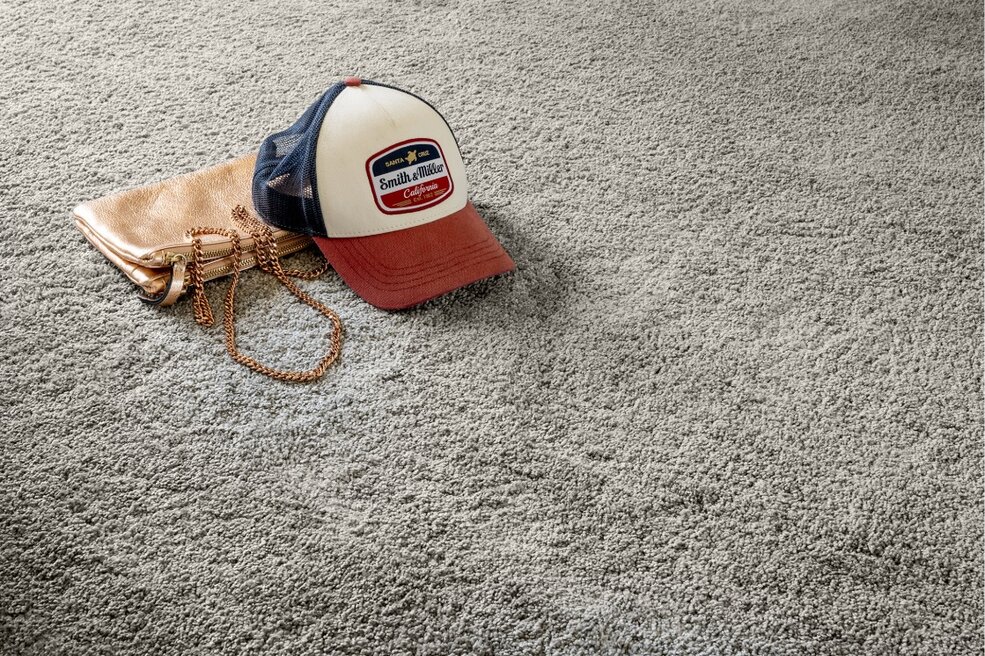 Grauer JOKA Teppich mit darauf liegender Baseballkappe und Accessoires.