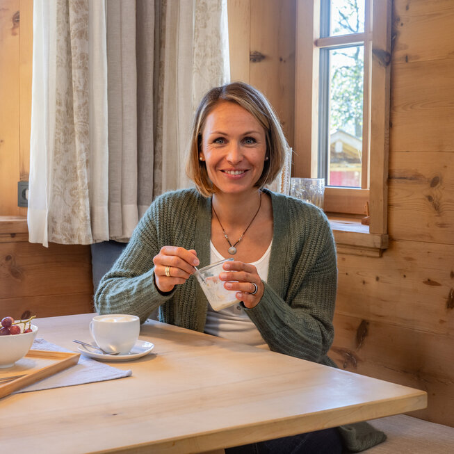 Magdalena Neuner am Tisch – Magdalena Neuner sitzt mit einer Tasse Tee an einem Esstisch, gemütliche Atmosphäre mit Holzelementen.