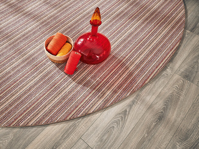 Nahaufnahme eines grauen JOKA Fußbodens, auf dem ein rot gemusterter Teppich liegt.