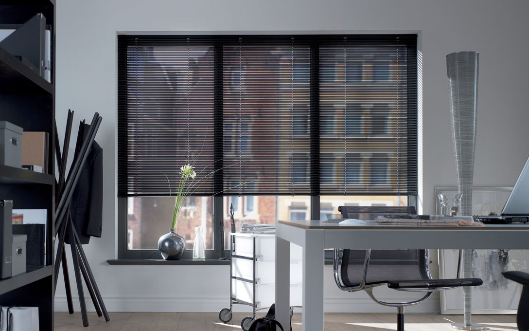 Ein Büroraum mit einer großen Fensterfront, vor der eine Jalousie aus Aluminium als Sonnenschutz hängt.