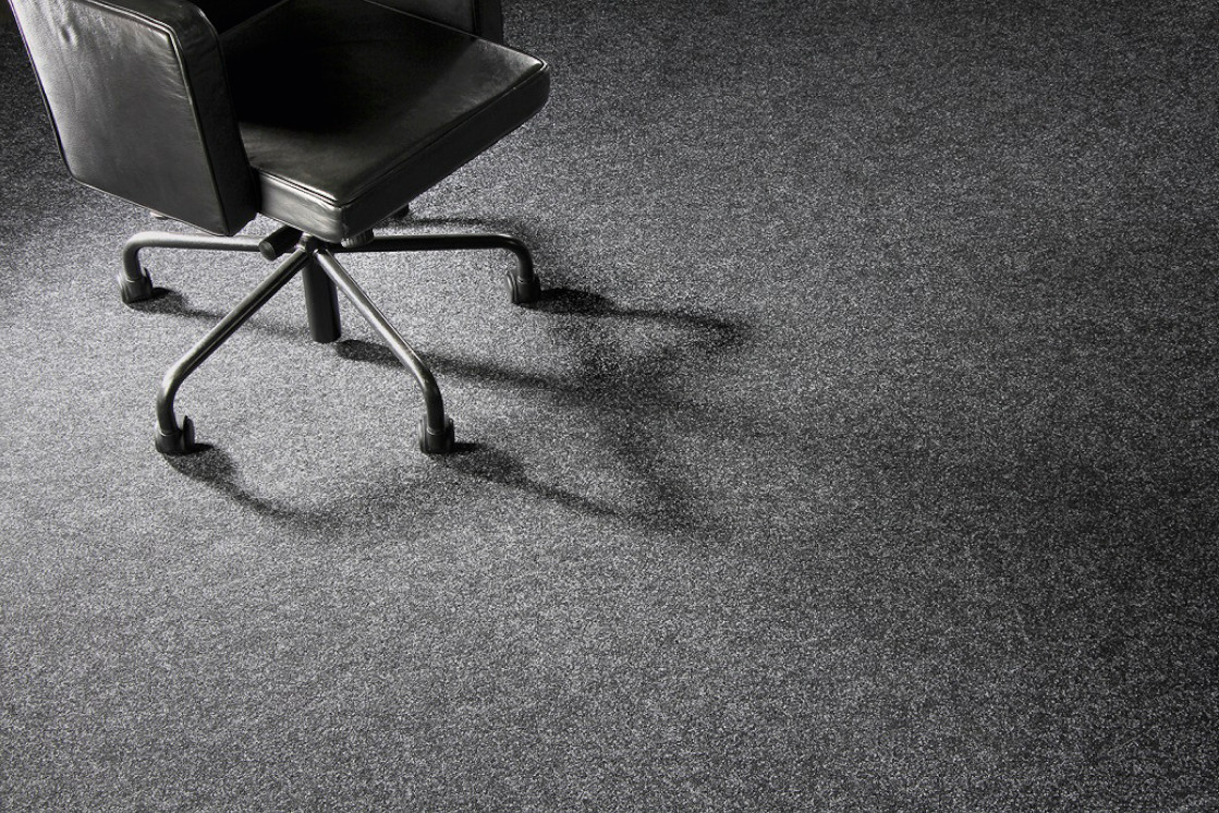 Ein schwarzer Bürostuhl mit Rollen steht auf einem dunkelgrauen Teppichboden und wirft einen Schatten, der die Struktur des JOKA Teppichs betont.