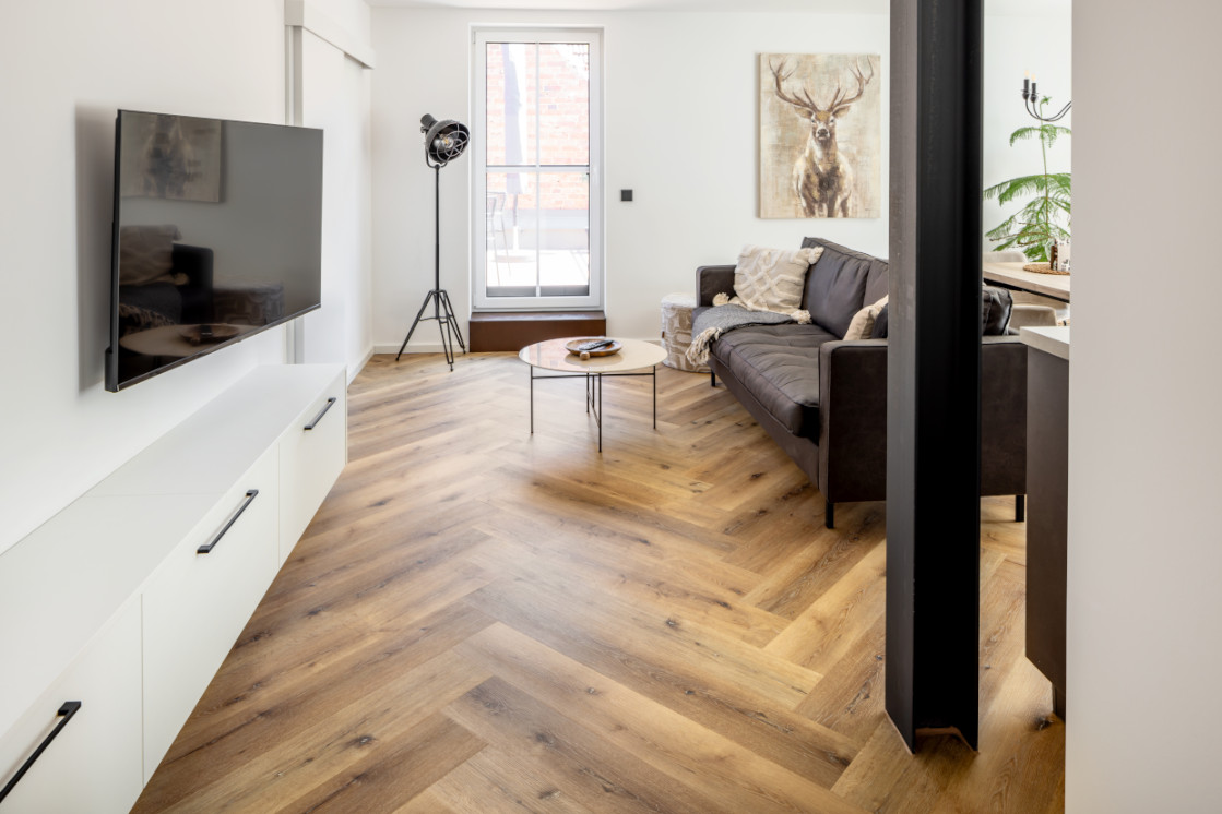 Modernes Wohnzimmer in einem Ferienhaus mit hellen Wänden, Parkettboden in Fischgrätoptik und offenem Übergang zur Küche.