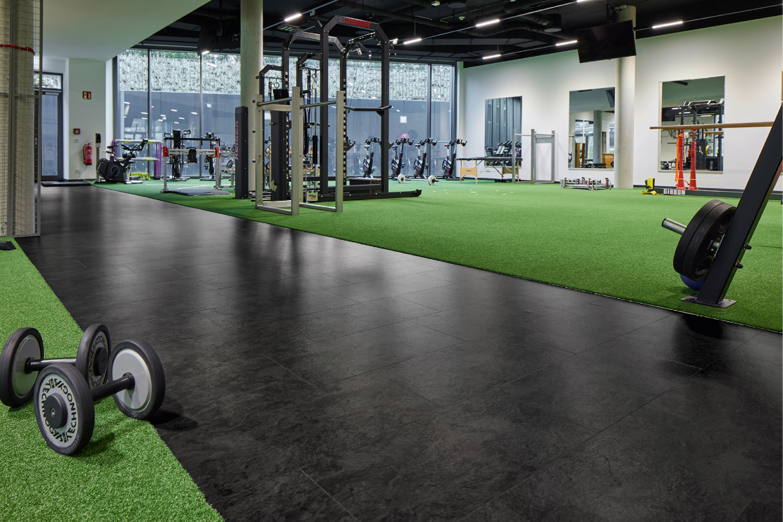Raumbild eines Fitnessstudios mit dunklem Designboden und grünem Teppich, auf welchem die Geräte stehen.