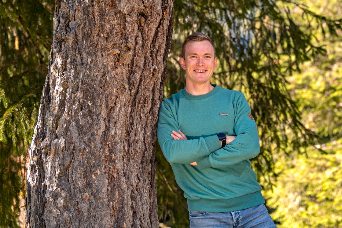 Benedikt lehnt entspannt mit verschränkten Armen an einem dicken Baum.