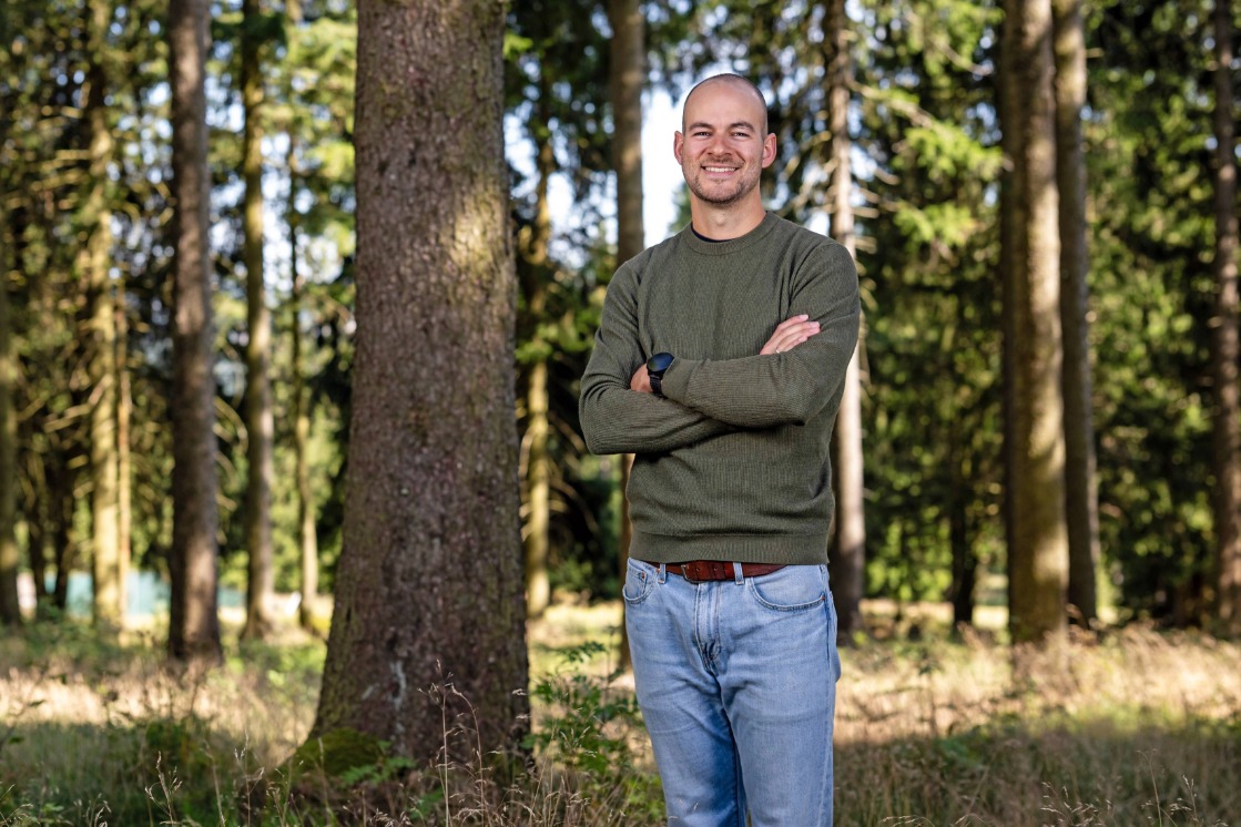 Danilo Riethmueller steht mit verschränkten Armen in einem Waldgebiet, umgeben von hohen Bäumen. Er trägt einen grünen Pullover, Jeans und eine Armbanduhr und lächelt freundlich in die Kamera.