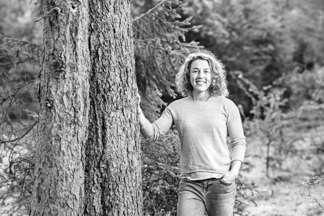 Ein schwarz-weiß Foto von Laura die lässig an einem Baum lehnt, umgeben von einer dichten, grünen Waldkulisse.